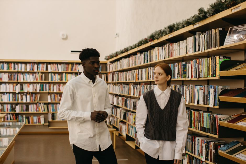 Two diverse students in white shirts converse in a cozy library setting, surrounded by books.