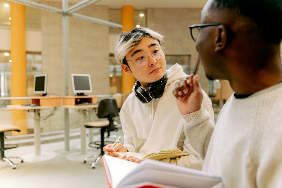 Two men discussing and holding notebooks in a library setting, engaging with each other.